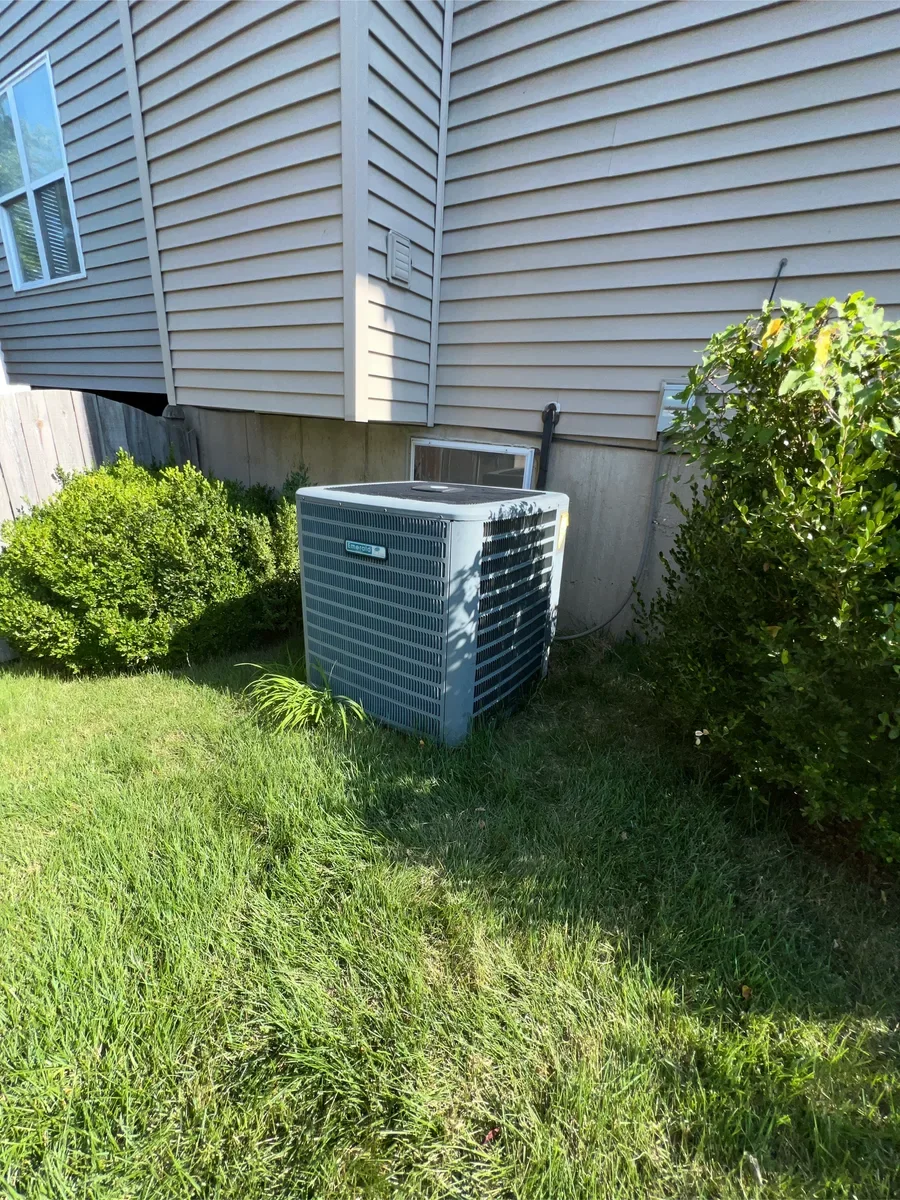 Original air conditioner between the basement window and shrubs before replacement in St. Peters.